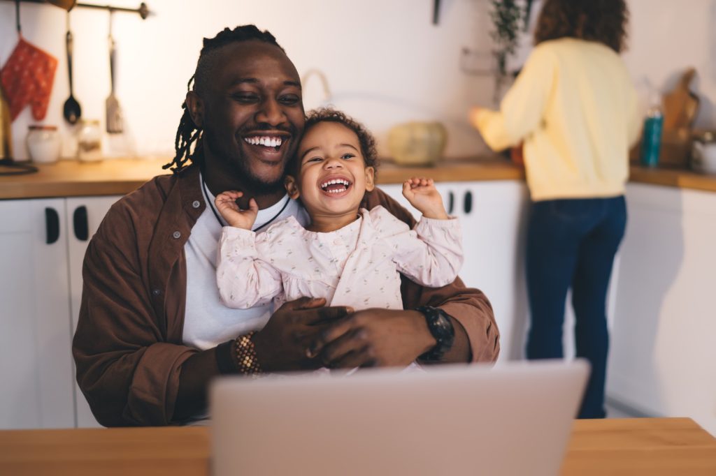 Cheerful black father and daughter laughing while sitting together at table with laptop