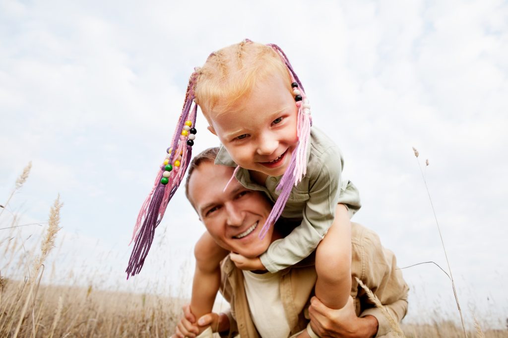 Portrait of happy red hair child sitting on shoulders father, smiling to camera. Laughing family