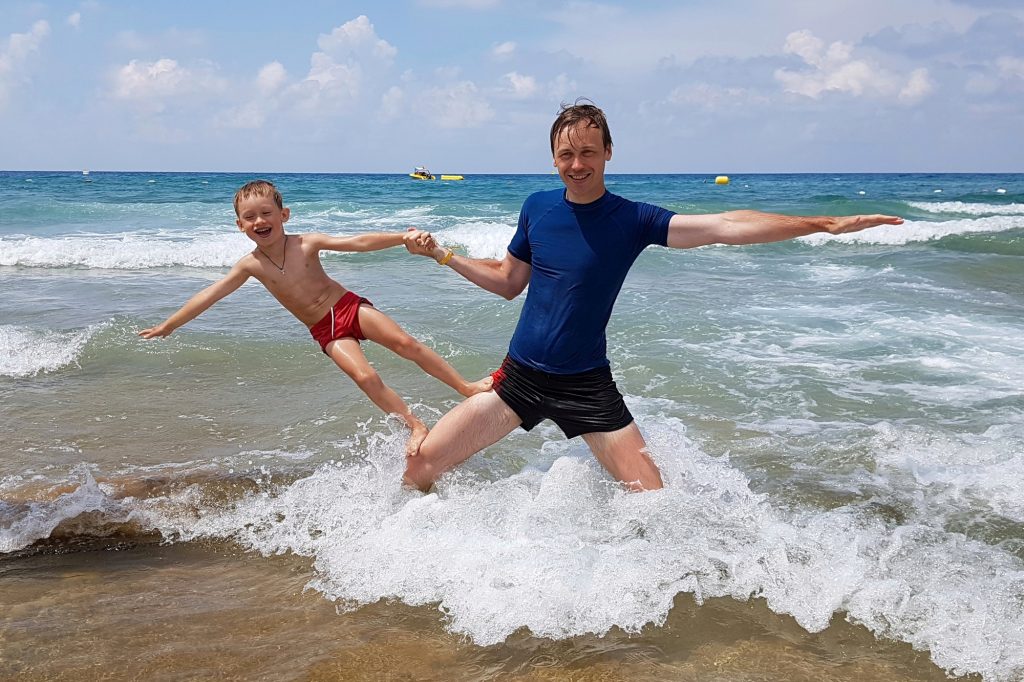 The father, standing on the beach in the water, put his son on his knee and holds his hand.